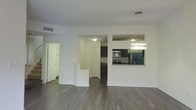 This thoughtfully designed two-floor space offers a clean, modern aesthetic throughout. The main living area features wide-plank, hardwood-style flooring that extends across the open-concept layout, creating a cohesive and inviting atmosphere. Crisp white walls provide a bright, neutral backdrop that enhances the sense of openness and space.

The kitchen is accessible via a pass-through window from the main living area, allowing for easy interaction between spaces. It is equipped with stainless steel appliances, including a refrigerator, oven, and microwave, complemented by dark cabinetry that adds a striking contrast to the light surroundings. A stylish pendant light fixture hangs above, providing both functional and ambient illumination.

A staircase with a white half-wall partition connects the two floors, adding architectural interest while maintaining an open feel. Adjacent to the staircase, a doorway leads to additional rooms, offering a well-organized and functional floor plan. The space is well-lit throughout, with ceiling-mounted light fixtures ensuring consistent brightness in every area.

Overall, this space balances contemporary design with practical functionality, making it suitable for a variety of uses. The combination of modern finishes, an open layout, and multi-floor configuration provides both comfort and versatility.