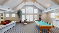 This single-floor community clubhouse is thoughtfully designed to offer a variety of social and recreational spaces under one roof. The centerpiece of the space is a dramatic vaulted game room, where soaring cathedral ceilings rise above a large-format arched window wall that floods the room with abundant natural light. A full-size pool table with a classic wood finish anchors the space, complemented by a patterned carpet, a small seating area, and a decorative indoor tree that adds a touch of organic warmth. Adjacent to the game room is a refined lounge area featuring an elegant coffered ceiling with recessed lighting and crisp white trim. Built-in white cabinetry with powder-blue accent backing frames a central fireplace and wall-mounted television, creating a welcoming focal point. Comfortable seating, including a tufted sofa and cognac leather accent chairs, is arranged to encourage relaxed conversation. The kitchen is outfitted with dark granite countertops, a central island with an undermount sink, stainless steel appliances, white cabinetry, and warm hardwood flooring. Soft blue walls carry a consistent, calming color palette throughout the entire space, contributing to a cohesive and inviting ambiance overall.