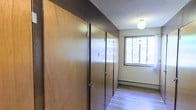 This single-floor space is defined by a prominent wall of floor-to-ceiling built-in storage units constructed from wood-panel doors in a warm brown finish. The units span nearly the full length of one wall, offering substantial organizational capacity. Each door is fitted with functional hardware, including latches and hinges, suggesting durable, utilitarian construction.

The flooring throughout is a light-toned hardwood-style material that contrasts cleanly with the darker wood tones of the storage wall. A textured popcorn ceiling adds a period-characteristic finish overhead, complemented by a simple globe light fixture that provides ambient illumination.

Along the opposite wall, a baseboard heater runs beneath a window that allows natural light to filter into the space, brightening the otherwise neutral interior. The window also offers a partial view of surrounding greenery, introducing a subtle connection to the outdoors.

The overall ambiance is functional and understated, with the built-in storage serving as the dominant architectural feature. The space balances practicality with a modest, no-frills aesthetic typical of mid-century residential construction.
