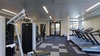 This fitness center offers a comprehensive workout environment designed to accommodate a range of exercise routines. The space is finished with a distinctive checkered carpet pattern in shades of gray and beige, adding visual interest to the floor while providing a functional surface underfoot. Overhead, recessed linear LED lighting illuminates the room evenly, complementing the natural light that flows in through large windows along one wall, offering views of an outdoor area beyond.

The strength training section is equipped with a cable machine and various resistance apparatuses, providing users with multiple options for targeted muscle work. On the opposite side of the room, a row of treadmills lines the windowed wall, allowing cardio users to enjoy the outdoor view during their workouts. The neutral color palette of white and gray walls contributes to a clean, modern aesthetic throughout the space.

A glass-paneled entry door with an illuminated emergency exit sign above it provides clear access and safety compliance. The overall layout is open and well-organized, ensuring ease of movement between equipment stations.