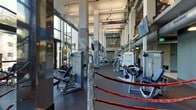 This modern fitness center is designed to accommodate a full range of workout needs within a spacious, open-plan layout. The facility features soaring ceilings with exposed ductwork and industrial-style finishes, giving the space an energetic and motivating atmosphere. Large floor-to-ceiling windows line the perimeter, flooding the interior with natural light and creating an airy, open feel.

The gym floor is outfitted with a comprehensive selection of strength-training machines and cable equipment, strategically arranged to allow for ease of movement between stations. A dedicated cardio zone offers multiple machines, including treadmills and seated cardio units, positioned beneath wall-mounted flat-screen televisions for an engaging workout experience. A standout feature of the facility is the full boxing ring, complete with ropes, providing a dedicated space for combat sports training and high-intensity workouts.

Reflective wall mirrors and textured metal panel accents contribute to the industrial aesthetic, while rubber flooring throughout ensures safety and durability. Structural columns punctuate the space, adding to its architectural character. Visible shelving units in the background suggest additional amenities or storage areas, rounding out this comprehensive and thoughtfully designed fitness facility.