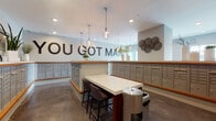 This thoughtfully designed mail room serves as a functional yet stylish communal hub. The space is defined by an extensive array of numbered individual mailboxes finished in a sleek gray tone with warm wood-trimmed ledges that wrap around the room's perimeter, providing ample mail storage for a large number of residents. At the center of the room, a white quartz-topped communal table with dark bar-height seating offers a convenient spot to sort through correspondence. The room's character is elevated by a bold "YOU GOT MAIL" wall installation, adding a lighthearted and welcoming personality to an otherwise utilitarian space. Industrial-style Edison bulb pendant lights hang from the white ceiling, complementing the polished concrete floors below and contributing to a contemporary aesthetic. A wall-mounted flat-screen display provides an informational focal point, while decorative elements — including woven wall art, ceramic vases with branches, and potted greenery — bring warmth and visual interest throughout. Interior windows connect the mail room to adjacent spaces, enhancing the sense of openness. Recessed lighting and natural light from glass-paneled entry doors ensure the space remains bright and inviting.