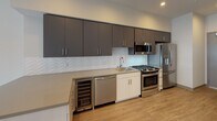Located at 11920 W Pico Blvd in the vibrant West Los Angeles neighborhood of 90064, this modern single-floor unit offers a thoughtfully designed layout with clean finishes throughout. The open-concept kitchen is a standout feature, equipped with stainless steel appliances including a gas range, microwave, dishwasher, refrigerator, and a built-in wine cooler. Charcoal gray upper cabinetry contrasts elegantly with lighter lower cabinets, complemented by a herringbone-patterned white tile backsplash and light quartz countertops. Light wood-look flooring runs seamlessly from the kitchen into the open living area, which benefits from natural light streaming through large windows. Recessed lighting throughout provides consistent, modern illumination. The bedroom includes a well-organized walk-in closet with built-in shelving and its own recessed light, offering ample storage. The bathroom features a clean, all-white tiled tub and shower combination with chrome fixtures, maintaining the unit's contemporary and minimalist aesthetic. The overall design is cohesive, with a neutral color palette and modern hardware choices that create a bright, functional, and stylish living environment.