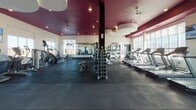 Located in Draper, Utah, this single-floor fitness center combines energetic design with comprehensive workout amenities. The entrance corridor immediately sets the tone with a bold, large-scale urban mural covering an entire wall, depicting a vivid cityscape rendered in striking reds, oranges, and blues. Geometric blue-and-charcoal patterned carpet tiles line the entryway, leading visitors into the main workout floor.

The primary exercise area features rubber flooring, floor-to-ceiling mirrors along the walls, and a dramatic deep-magenta ceiling accented by recessed lighting and ceiling fans. This space is well-stocked with cardio machines including treadmills and stair climbers, a full dumbbell rack, and cable weight machines. Large windows allow abundant natural light to fill the room.

A glass-enclosed children's play area is seamlessly integrated into the facility, featuring colorful checkerboard flooring, whimsical tree murals, bright toys, and a small table—providing a safe, cheerful space for young visitors.

A separate studio room offers an open, flexible space for spin cycling, yoga, or group fitness. Outfitted with rubber flooring, a spin bike, yoga mats, and colorful wall art, the room is bathed in natural light through oversized windows that frame sweeping views of the surrounding Utah landscape and distant mountains.
