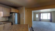 This single-floor space features an open layout that connects the kitchen to a carpeted living area, creating a functional and cohesive flow throughout. The kitchen is equipped with a stainless steel refrigerator and upper cabinetry in a warm wood finish, complemented by granite countertops that add a touch of durability and style. The warm tan walls carry consistently through both the kitchen and adjoining living space, contributing to a unified and neutral aesthetic. A structural column subtly defines the transition between the two areas without fully separating them. The living area benefits from natural light streaming in through two windows flanking a paneled entry door, which is framed by dark curtains that offer contrast against the lighter walls. Carpeting in the living space adds comfort underfoot. Smoke detectors are visible on the ceiling, and standard electrical outlets are positioned along the walls. The overall ambiance is practical and straightforward, with a clean, move-in-ready feel suited for everyday living.