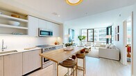 This well-appointed 2-bedroom, 2-bathroom residence is designed with a contemporary aesthetic, featuring clean lines, a neutral palette, and light wood flooring that flows seamlessly throughout the single-story layout. The open-concept living, dining, and kitchen area serves as the heart of the home, anchored by a striking wood-topped island with bar seating. The kitchen is outfitted with stainless steel appliances, flat-front light wood cabinetry, open shelving, and a crisp white countertop, offering both style and functionality. The living room is bathed in natural light through expansive floor-to-ceiling windows, providing generous views of the surrounding cityscape and creating an airy, inviting atmosphere. The primary bedroom is a spacious retreat with large windows framing urban views, while the second bedroom is a comfortable, well-proportioned room with warm ambient lighting. Both bathrooms are finished in a minimalist style, featuring light wood vanities, large-format tile flooring, and clean white fixtures. A dedicated closet, hallway, and thoughtful storage solutions round out the practical layout, making this residence as functional as it is visually refined.
