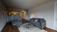 This single-floor space offers a thoughtfully designed open-concept layout that seamlessly connects the living area and kitchen, creating a cohesive and functional environment. The living room is anchored by rich, dark hardwood flooring and features plush gray upholstered seating, including a full sofa and accent chair, arranged around a rustic round coffee table atop a patterned area rug. Neutral gray walls provide a calm, versatile backdrop that enhances the overall sense of warmth and comfort. A table lamp adds soft ambient lighting to complement the room's inviting atmosphere. The kitchen is positioned at the rear of the open floor plan and is equipped with stainless steel appliances, including a refrigerator and microwave, alongside white cabinetry and a peninsula counter with bar seating — ideal for casual dining or entertaining. Track lighting illuminates the kitchen workspace effectively. A hallway off the main living area leads to additional rooms, with a classic paneled door and a wall clock adding subtle decorative touches. The consistent hardwood flooring flows throughout the space, tying the rooms together and reinforcing a clean, modern aesthetic. Overall, this space offers a practical and stylish single-level layout well-suited for comfortable everyday living.