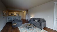 This single-floor space offers a thoughtfully designed open-concept layout that seamlessly connects the living area and kitchen, creating a cohesive and functional environment. The living room is anchored by rich, dark hardwood flooring and features plush gray upholstered seating, including a full sofa and accent chair, arranged around a rustic round coffee table atop a patterned area rug. Neutral gray walls provide a calm, versatile backdrop that enhances the overall sense of warmth and comfort. A table lamp adds soft ambient lighting to complement the room's inviting atmosphere. The kitchen is positioned at the rear of the open floor plan and is equipped with stainless steel appliances, including a refrigerator and microwave, alongside white cabinetry and a peninsula counter with bar seating — ideal for casual dining or entertaining. Track lighting illuminates the kitchen workspace effectively. A hallway off the main living area leads to additional rooms, with a classic paneled door and a wall clock adding subtle decorative touches. The consistent hardwood flooring flows throughout the space, tying the rooms together and reinforcing a clean, modern aesthetic. Overall, this space offers a practical and stylish single-level layout well-suited for comfortable everyday living.