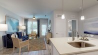 This single-story residence offers a thoughtfully designed open-concept floor plan that seamlessly connects the kitchen, dining, and living areas. The kitchen is outfitted with white shaker cabinetry, quartz countertops, stainless steel appliances, and a central island with pendant lighting and bar seating—ideal for both cooking and casual entertaining. Light wood-look flooring extends throughout the main living areas, contributing to a clean, cohesive aesthetic. The dining area accommodates a glass-top table with seating for four, accented by a decorative mirror and gallery wall. The living room features large windows that allow ample natural light, complemented by a ceiling fan and neutral gray walls.

The primary bedroom is carpeted and generously sized, with natural light filtering through blinds and drape-framed windows. A second bedroom also features carpet, a ceiling fan, and window treatments. The bathroom includes a glass-enclosed shower, light wood-look flooring, white cabinetry, and a quartz vanity top. A dedicated laundry area houses a full-size washer and dryer with wire shelving for storage. Overall, the space presents a modern, neutral palette throughout.
