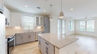 This well-designed 1-bedroom, 1-bathroom residence offers a clean, contemporary aesthetic across a single floor. The open-concept kitchen and living area serves as the heart of the unit, anchored by a large central island with a striking quartz countertop and pendant lighting above. The kitchen features two-tone cabinetry — soft gray shaker-style lower cabinets paired with white upper cabinets, including glass-front display sections — complemented by stainless steel appliances and a subway tile backsplash. Light wood-look flooring runs seamlessly throughout the entire space, enhancing the airy, cohesive feel. The living area benefits from abundant natural light through multiple large windows and a glass door that opens to a balcony with outdoor views. The bedroom is generously proportioned, offering dual windows with outdoor views, a ceiling fan, and direct access to a walk-in closet equipped with built-in shelving and hanging rods for ample storage. The bathroom, visible from the hallway, features tiled finishes. Recessed lighting and crisp white walls contribute to the bright, modern atmosphere that defines this thoughtfully laid-out residence.