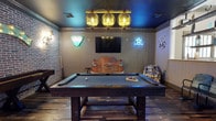 This game room is a thoughtfully designed entertainment space that blends rustic charm with bold, playful character. The centerpiece is a full-sized pool table with a dark wood base and gray felt surface, positioned beneath a striking industrial-style chandelier featuring three cage-pendant lights that cast a warm, atmospheric glow across the room.

The walls are adorned with eye-catching neon signs, including a Dallas Cowboys star emblem and a colorful billiards-themed sign, adding vibrant splashes of color to the space. A Texas Route 66 sign reinforces the room's Americana theme. A wall-mounted flat-screen TV is prominently displayed, making this space equally suited for watching games or movies.

One of the room's standout decorative elements is a vintage Chevrolet truck front repurposed as a unique bench or seating piece, adding a one-of-a-kind rustic focal point. The flooring features wood-look planks that complement the room's warm, laid-back aesthetic, while exposed brick-style wall accents add texture and depth. The dark painted ceiling enhances the intimate, lounge-like ambiance, making this game room a welcoming and lively gathering space.