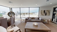 This contemporary single-floor space is defined by its clean lines, neutral palette, and an abundance of natural light. The open-plan living area is an immediate standout, anchored by large floor-to-ceiling windows that frame sweeping panoramic views of the surrounding cityscape and sky beyond. The lounge area features a plush taupe sofa, a substantial natural wood coffee table, and warm woven area rugs that add texture to the light-toned flooring. Curated artwork, decorative plants, and organic accent pieces throughout lend the space a refined yet welcoming character. Recessed lighting and smooth white ceilings contribute to a bright, airy atmosphere. A glass-paneled display cabinet provides stylish storage along one wall, complementing the overall modern aesthetic. The kitchenette area is equally well-appointed, featuring sleek dark cabinetry with polished hardware, continuous white quartz countertops, and a textured wave-pattern tile backsplash. A wall-mounted flat-screen television, a beverage refrigerator, and dedicated countertop appliances make this area fully functional. A central island with bar-stool seating provides a casual gathering space. Wood-look tile flooring runs through the kitchen zone, adding warmth and cohesion. Together, the spaces offer a sophisticated, contemporary environment ideal for both relaxation and entertaining.