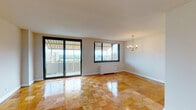 This well-appointed single-floor residence is unified by polished parquet wood flooring in a classic checkered pattern that extends throughout every room, lending warmth and continuity to the entire space. The generous living and dining area serves as the heart of the home, bathed in natural light from large sliding glass doors that open onto a private balcony. From here, expansive views of the surrounding cityscape and tree-lined greenery create a striking backdrop. An elegant brass chandelier adds a traditional accent to the dining area. The kitchen is outfitted with white cabinetry, dark granite-style countertops, a stainless steel sink, and a full-size refrigerator, providing a clean and functional workspace. The bedrooms are comfortably proportioned, each featuring large windows that frame elevated city and park views, along with individual climate control units. Closet space is well integrated throughout. The bathroom features classic white wall tiling, a combined tub and shower, and a mosaic tile floor with warm accent tones. Clean white walls and neutral finishes throughout the residence create a bright, airy ambiance, while the elevated position of the unit ensures privacy and remarkable panoramic views from nearly every room.