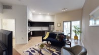 This single-floor clubhouse is designed with a sleek, contemporary aesthetic that promotes both comfort and socializing. The open-plan layout centers around a spacious lounge area furnished with a large, plush sectional sofa and a wooden coffee table accented with decorative succulents, creating a welcoming atmosphere for residents and guests alike.

At the heart of the space is a well-appointed wet bar, featuring dark cabinetry, a light-toned countertop, and bar stool seating — ideal for casual entertaining. A wall-mounted television is positioned above the bar, framed by a striking exposed brick accent wall that adds warmth and texture to the otherwise clean, minimalist interior. Pendant lighting in brushed gold hangs above the bar, providing focused illumination and a touch of elegance.

The space is finished with smooth white walls, recessed ceiling lighting, and wood-tone flooring that runs throughout, contributing to a cohesive and polished look. Tall indoor plants add a natural element to the design. Large windows allow natural light to filter in, enhancing the bright and airy feel of the room. Overall, the clubhouse offers a refined yet relaxed environment well-suited for community gatherings and everyday leisure.