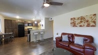 This single-floor space offers a thoughtfully designed open-concept layout that seamlessly connects the kitchen and living area. The living room features rich, dark hardwood-style flooring that extends throughout, grounding the space with warmth and continuity. A ceiling fan overhead provides both function and a finishing touch to the room.

The kitchen is well-appointed with dark cabinetry, black appliances including a refrigerator, microwave, and range, and a breakfast bar with modern white bar stools that serve as a natural divider between the cooking and living areas. Pendant lighting hangs above the bar, adding a contemporary accent to the kitchen zone.

The living area is open and inviting, offering ample room for comfortable seating. Textured ceilings add architectural character throughout the space, while neutral wall tones provide a versatile backdrop. A doorway visible from the main living area suggests additional rooms beyond, offering a sense of depth and privacy within the floor plan. Overall, the space balances practical design with a welcoming ambiance suited for everyday living.