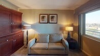 Unit #603 at 801 National City is a fully furnished, single-floor residence thoughtfully appointed for comfortable living. The bedroom serves as a restful retreat, centered around a queen-sized bed with a rich mahogany headboard, flanked by matching nightstands and warm table lamps. A full-length mirrored sliding closet adds depth to the room while maximizing storage. Framed artwork on soft neutral walls contributes to the room's calm, inviting atmosphere, and carpeted flooring adds warmth underfoot. The living area features a plush loveseat accented by coordinating floor and table lamps, creating a cozy, well-lit space ideal for relaxing. Matching framed artwork ties the aesthetic together seamlessly. The kitchen is equipped with granite countertops, light wood cabinetry, a built-in cooktop, and an overhead microwave, offering a functional and stylish cooking space. Tile flooring provides easy maintenance throughout the kitchen and bathrooms. The bathroom showcases granite countertops with an undermount sink, light wood vanity, a large wall mirror with overhead lighting, and tile flooring — delivering a clean and polished finish. Overall, the unit presents a cohesive, well-maintained interior with warm tones and practical furnishings throughout.