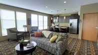 This single-story space features an open-concept layout that integrates the living room, dining area, and kitchen into one cohesive, functional environment. The living area is anchored by a patterned sofa dressed with colorful accent pillows and paired with a round coffee table, creating a comfortable and inviting gathering space. Large windows fitted with white blinds allow generous natural light to fill the room, enhancing the warm, neutral wall tones.

The kitchen is outfitted with dark espresso cabinetry, stainless steel appliances including a refrigerator and range, and a light-toned countertop that extends into a breakfast bar with seating. Pendant lights with warm amber shades hang above the kitchen area, adding a stylish focal point that complements the recessed ceiling lighting found throughout the space.

The dining area bridges the kitchen and living room, featuring a table and chairs that maintain the open flow of the layout. The multi-toned area rug grounds the living space and adds texture and depth. Overall, the space offers a modern, well-appointed design with a cohesive color palette and practical, open arrangement.
