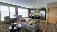 This single-story space features an open-concept layout that integrates the living room, dining area, and kitchen into one cohesive, functional environment. The living area is anchored by a patterned sofa dressed with colorful accent pillows and paired with a round coffee table, creating a comfortable and inviting gathering space. Large windows fitted with white blinds allow generous natural light to fill the room, enhancing the warm, neutral wall tones.

The kitchen is outfitted with dark espresso cabinetry, stainless steel appliances including a refrigerator and range, and a light-toned countertop that extends into a breakfast bar with seating. Pendant lights with warm amber shades hang above the kitchen area, adding a stylish focal point that complements the recessed ceiling lighting found throughout the space.

The dining area bridges the kitchen and living room, featuring a table and chairs that maintain the open flow of the layout. The multi-toned area rug grounds the living space and adds texture and depth. Overall, the space offers a modern, well-appointed design with a cohesive color palette and practical, open arrangement.