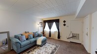 This single-floor residence offers a welcoming and functional layout designed for comfortable everyday living. The living room serves as the heart of the home, featuring wall-to-wall carpeting that adds warmth underfoot and a textured tile ceiling that contributes a classic, traditional character to the space. A large double window dressed with elegant dark curtains and sheer underlays allows natural light to filter softly into the room, creating a bright yet cozy ambiance. The space comfortably accommodates a sectional sofa in a calming blue-gray tone, accented with decorative pillows, and is anchored by a patterned area rug that adds visual interest to the floor. A floor lamp in the corner provides additional ambient lighting, while framed wall art and decorative diamond-shaped wall accents lend a personal, lived-in charm. A wall-mounted thermostat and a utility access panel near the entryway point to practical, well-integrated home systems. The entryway door with a screen insert adds to the home's functional design. Overall, the space presents a warm, understated aesthetic with a focus on comfort and practicality.