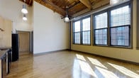 This 2-bedroom, 1-bathroom unit showcases a compelling blend of industrial and modern design elements throughout its single floor. The living area immediately draws attention with its soaring ceilings, where exposed wooden beams and visible ductwork pay homage to the building's industrial heritage. Large, multi-pane windows flood the space with generous natural light, casting warm reflections across the smooth hardwood floors below. The open layout of the main living area creates a spacious, airy atmosphere well-suited for both relaxing and entertaining. Pendant industrial-style lighting fixtures complement the raw architectural details overhead, reinforcing the loft aesthetic. The contrast between the crisp white walls and the warm, natural tones of the wood ceiling creates a visually dynamic environment. The hardwood flooring runs throughout, adding warmth and continuity to the space. The unit's design successfully combines the character of a converted industrial building with modern residential comfort, resulting in a distinctive living space with lasting architectural appeal.