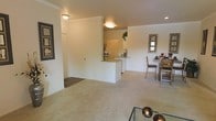 This single-story space is designed with an open-concept layout that seamlessly connects the living, dining, and kitchen areas into one cohesive environment. The living area features plush, neutral-toned carpeting that extends throughout the main spaces, contributing to a warm and comfortable atmosphere. Crown molding lines the upper edges of the walls, adding a refined architectural detail to the otherwise clean, minimalist interior.

A half-wall partition separates the kitchen from the living space, creating a sense of definition without completely closing off the areas from one another. The kitchen features a raised breakfast bar along the partition, offering a casual dining or prep surface. A full-size refrigerator is visible within the kitchen, and recessed lighting supplements the flush-mount ceiling fixtures found throughout the space.

Adjacent to the kitchen, the dining area accommodates a dining table and chairs beneath a decorative mirror, creating a functional yet stylish space for meals. The overall color palette relies on soft whites and warm beiges, lending the interior a bright and neutral character. The layout is practical and well-organized, making efficient use of the available space across its single floor.