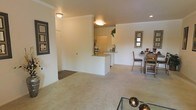 This single-story space is designed with an open-concept layout that seamlessly connects the living, dining, and kitchen areas into one cohesive environment. The living area features plush, neutral-toned carpeting that extends throughout the main spaces, contributing to a warm and comfortable atmosphere. Crown molding lines the upper edges of the walls, adding a refined architectural detail to the otherwise clean, minimalist interior.

A half-wall partition separates the kitchen from the living space, creating a sense of definition without completely closing off the areas from one another. The kitchen features a raised breakfast bar along the partition, offering a casual dining or prep surface. A full-size refrigerator is visible within the kitchen, and recessed lighting supplements the flush-mount ceiling fixtures found throughout the space.

Adjacent to the kitchen, the dining area accommodates a dining table and chairs beneath a decorative mirror, creating a functional yet stylish space for meals. The overall color palette relies on soft whites and warm beiges, lending the interior a bright and neutral character. The layout is practical and well-organized, making efficient use of the available space across its single floor.