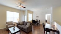 This single-story residence features an open-concept layout that seamlessly connects the living and dining areas, creating a spacious and functional environment. The living area is anchored by a comfortable gray sofa accented with patterned throw pillows, complemented by a side table with a classic table lamp. A ceiling fan with a built-in light fixture overhead provides both comfort and ambient lighting.

The warm hardwood flooring flows throughout the space, adding a cohesive and inviting feel. Neutral cream-toned walls enhance the brightness, aided by several large windows fitted with white blinds that allow generous natural light to fill the room.

Transitioning into the dining area, a dark wood dining table surrounded by matching chairs sits beneath a pendant light fixture, adding a refined focal point to the space. Decorative wall accents add a subtle artistic touch nearby.

Beyond the main living areas, a hallway leads further into the residence, hinting at additional rooms. The overall ambiance is clean, bright, and welcoming, with a practical open layout that promotes easy movement and a comfortable everyday living experience.
