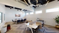 This thoughtfully designed single-floor office space seamlessly blends industrial and contemporary aesthetics to create a productive and visually appealing work environment. The space is defined by its striking exposed black ceiling with visible ductwork and linear LED lighting, which contrasts beautifully against the crisp white walls and whitewashed brick wainscoting that runs throughout.

The open coworking area features modular white workstations with privacy dividers and ergonomic chairs, offering flexible seating for multiple occupants. A herringbone-patterned carpet tile floor adds a refined, textural element underfoot. High clerestory windows line the upper walls, drawing in natural light while maintaining privacy, and the generous ceiling height contributes to an airy, open atmosphere.

A dedicated conference area accommodates a large rectangular table surrounded by navy blue upholstered chairs with gold accents, anchored by a striking globe chandelier overhead. A vibrant tropical-themed artwork adorns the accent wall, adding a warm focal point to the meeting space. Interior glass partitions allow visual connectivity between zones while preserving a sense of structure. The space is fully equipped with office essentials including a printer station, making it a functional and well-appointed professional environment.