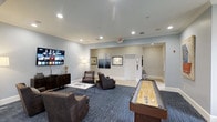 This single-floor community lounge is designed to offer both comfort and entertainment in a well-appointed setting. The space features a contemporary aesthetic, with soft neutral-toned walls, recessed lighting, and clean crown molding that lend a polished, inviting atmosphere throughout.

The main lounge area is anchored by a large wall-mounted smart TV, complemented by a dark wood media console. Comfortable seating — including plush armchairs and a sofa arranged around a sleek glass coffee table — sits atop a rich blue area rug, creating a cozy gathering spot ideal for socializing or unwinding.

A standout feature of the space is the full-size shuffleboard table, positioned prominently to one side, offering a fun recreational element that enhances the lounge's communal appeal. Decorative wall art and a floor lamp add warmth and personality to the room.

Beyond the main lounge, a connected corridor leads to additional areas, with tile flooring and recessed lighting continuing the clean, modern design language. The layout flows naturally between zones, making the space functional for a variety of uses. Overall, the lounge strikes a balance between a relaxed residential feel and an engaging, activity-oriented environment suited for community gatherings.