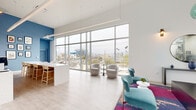 Located at 5401 Lankershim Blvd in North Hollywood, this contemporary amenity space serves as a stylish communal hub designed with both comfort and functionality in mind. The single-floor layout is anchored by an expansive lounge area distinguished by soaring ceilings and dramatic floor-to-ceiling windows that frame sweeping views of the surrounding Los Angeles skyline and palm-lined streetscape, flooding the interior with abundant natural light.

The lounge is thoughtfully furnished with teal velvet accent chairs, plush swivel seating, and vibrant multicolored area rugs that add a bold pop of color against the light hardwood-style flooring and crisp white walls. A bold blue accent wall serves as a striking focal point, adorned with curated wall art and a decorative round mirror, while pendant lighting in aqua glass tones adds an artistic touch overhead.

A kitchenette area features sleek blue cabinetry, white quartz countertops, a geometric tile backsplash, and a gallery wall of framed artwork, providing a welcoming space for residents to gather. The space connects seamlessly to an outdoor pool courtyard, where lounge chairs, market string lights, shade umbrellas, and lush landscaping create a resort-like retreat within the heart of the urban community.