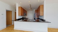This well-appointed urban residence offers a thoughtfully designed single-floor layout that balances open living spaces with private retreats. The living area is a standout, featuring expansive floor-to-ceiling windows that flood the space with natural light and frame impressive panoramic city views, including neighboring architecture and skyline landmarks. Light hardwood flooring runs throughout the main living area, enhancing the bright, airy atmosphere.

The open-concept kitchen is both functional and stylish, boasting rich warm-toned wood cabinetry, dark granite countertops, stainless steel appliances including a gas range with microwave above, and a refrigerator. A peninsula with an integrated sink and additional counter seating seamlessly connects the kitchen to the living space. Track lighting overhead adds a contemporary touch.

The primary bedroom is generously sized with plush carpet flooring, and includes access to a spacious walk-in closet outfitted with multi-tiered wire shelving systems for ample storage. Its en-suite bathroom features a granite-topped vanity with warm wood cabinetry, a glass-enclosed walk-in shower, and tile flooring. A second bedroom also enjoys large windows with city views and a dedicated closet. The second full bathroom offers a granite vanity and a full soaking tub with shower combination.