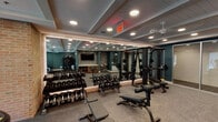 This single-floor fitness center is thoughtfully designed to support a wide range of workout routines in a polished, professional environment. The space is anchored by a spacious main workout floor with coffered ceilings, recessed lighting, and ceiling fans that keep the area comfortable during intense sessions. Exposed brick accent walls add a warm, industrial character that contrasts beautifully with the deep teal painted walls found throughout.

The primary exercise area is extensively equipped with multiple racks of dumbbells and kettlebells, a cable machine and power rack system, adjustable weight benches, and a row of cardio machines including treadmills and an elliptical. Full-length mirrors line the walls, visually expanding the space and allowing users to monitor their form. A wall-mounted flat-screen television provides entertainment during workouts.

A separate room offers cardio-focused equipment such as a stair climber and rowing machine, with large windows and roller shades that invite natural light and views of the surrounding exterior.

An additional mirrored studio room features a ballet barre along a brick accent wall, suitable for stretching, yoga, or dance-based fitness activities. Ceiling fans and recessed lighting maintain consistent comfort throughout.

Near the entrance, a dual-level hydration station with a bottle-filling option is set against a striking textured tile wall, ensuring convenient access to water for all users.