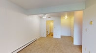 This single-floor residence offers a straightforward, functional layout with a clean and neutral aesthetic throughout. The main living area is finished with wall-to-wall carpeting and features a ceiling fan with integrated lighting, a baseboard heating system, and panel-style wall detailing. The open sightline from the living area into the kitchen makes the space feel connected and practical.

The galley-style kitchen is equipped with warm wood-toned cabinetry — both upper and lower — paired with a white countertop, a single-basin sink, a white refrigerator, and a range. The tile flooring in the kitchen provides a clean contrast to the carpeted living spaces, and a doorway leads further into the unit for easy flow.

The bathroom includes a full tub and shower combination with tiled surround, a wood-vanity with a white countertop sink, a wall-mounted medicine cabinet mirror, and a lighted vanity bar above — offering a bright and functional space.

A secondary carpeted room provides a versatile space with a large window dressed in vertical blinds and a baseboard heater beneath it. Another room features a sliding glass door that opens directly to a patio with views of a well-maintained lawn, allowing natural light to fill the space and offering a seamless connection to the outdoors.