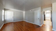 This single-story home in Dallas, Texas offers a thoughtfully designed layout comprising a living room, kitchen, bedroom, bathroom, laundry area, closet, and hallway. The living room is a standout space featuring rich, warm-toned hardwood floors, neutral gray walls, and clean white trim that together create a bright and inviting atmosphere. The open sightlines connect seamlessly to the hallway and kitchen beyond. The kitchen is a functional galley-style space finished with large-format tile flooring, dark cabinetry with open shelving towers on either side, and stainless steel appliances including a refrigerator. Natural light filters in through a window near the exterior door. The bedroom is a comfortable, well-proportioned room with plush carpeting, a ceiling fan with light fixture, and a window with blinds that brings in natural light. The full bathroom is well-appointed with tile flooring, a dark vanity topped with granite countertops, matte black fixtures, a large mirror, and a tub/shower combination. A dedicated laundry area accommodates a full-size washer and dryer. Outside, the brick exterior leads to a private yard with maintained grass and a wooden privacy fence, offering a pleasant outdoor retreat.
