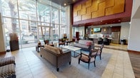 This thoughtfully designed clubhouse delivers an upscale communal experience from the moment you enter. The grand lobby immediately commands attention with soaring double-height ceilings and an impressive floor-to-ceiling glass curtain wall that frames sweeping views of the surrounding palm tree-lined courtyard and pool area, flooding the space with abundant natural light. Decorative wood-paneled accent walls in warm honey tones add architectural texture and visual warmth to the interior.

The main lounge area is furnished with plush sofas, upholstered armchairs, and stylish coffee tables arranged on area rugs, creating an inviting social atmosphere. Tucked beside the expansive windows, a cozy nook features bold, high-backed red patterned lounge chairs paired with a small side table—an intimate retreat with scenic outdoor views.

The fully equipped kitchen and entertaining area is a standout feature, boasting rich dark wood cabinetry, light quartz countertops, a mosaic tile backsplash, and stainless steel appliances including a full-size refrigerator. A large central island with bar seating and dramatic pendant lighting makes it ideal for gatherings and events. A wall-mounted flat-screen TV completes the entertainment setup.

Additional corridors connect to secondary seating areas, with a mix of tile and wood-look flooring transitions adding visual contrast throughout. The overall ambiance is modern, warm, and polished.
