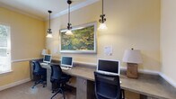 This single-floor business center and lounge offers a thoughtfully designed space that balances productivity with comfort. The room is finished with warm yellow walls, crisp white crown molding, and neutral carpeting that together create a welcoming, professional atmosphere.

A built-in concrete workstation counter runs along one wall, accommodating multiple computer workstations paired with ergonomic task chairs. Elegant wrought-iron pendant lights hang above the desk area, providing focused task lighting, while a framed impressionist-style water lily painting serves as a central decorative focal point above the workstations.

The lounge area features plush dark sofas and loveseats arranged around a round wooden coffee table, offering a relaxed setting ideal for casual meetings or reading. Framed artwork decorates the walls throughout, contributing to the refined aesthetic of the space.

A secondary area houses printing and copying equipment on a dark wood console, alongside frosted glass interior windows that allow light to filter between areas while maintaining privacy. Recessed ceiling lights and a ceiling fan provide additional illumination and climate comfort. A corridor with French doors connects to adjacent spaces, enhancing the overall flow and accessibility of the facility.