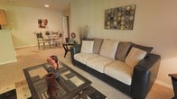 This single-floor space offers a comfortable and cohesive open-concept layout, where the living area flows naturally into an adjoining dining space. The living room is anchored by a generously sized sofa with dark leather trim and plush beige cushioning, complemented by a glass-top coffee table with a dark wood frame that adds a contemporary touch. A framed mixed-media artwork adorns the wall, adding visual interest and color to the otherwise neutral palette of warm beige and cream tones. Decorative accents, including dried botanical arrangements and candle holders, contribute to the room's inviting atmosphere. Soft, wall-to-wall carpeting runs throughout, enhancing the sense of warmth and comfort underfoot. The dining area, visible in the background, features a compact bistro-style table paired with ornate metal chairs, making efficient use of the space. Recessed lighting provides ambient illumination, and the open doorway between rooms promotes an easy sense of flow and connectivity. Overall, the space presents a well-coordinated, functional interior with a neutral, earthy color scheme suited to relaxed, everyday living.