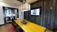 This single-story space in Flower Mound showcases a well-appointed meeting or gathering room with a distinctive rustic-modern aesthetic. The centerpiece is a substantial warm-toned wood communal table surrounded by woven metal chairs, anchored by a bold geometric pendant light fixture overhead. A dark-painted ceiling adds drama and contrast to the lighter surrounding walls.

A striking reclaimed wood accent wall serves as a focal point, housing a wall-mounted flat-screen television and providing rich textural depth to the room. The flooring transitions between light hardwood and brick-toned tile, adding visual interest throughout. A patterned area rug in red tones adds a pop of color beneath the seating area.

Large floor-to-ceiling windows dressed with sheer linen curtains flood the space with natural light and offer views of the surrounding streetscape. The room is accessed through elegant black-framed glass double doors, which lend a refined, professional touch and maintain visual connectivity to adjacent areas. Decorative elements, including framed artwork and an iron display easel, contribute to the room's curated, welcoming character. The overall ambiance balances functionality with distinctive style, making it well-suited for gatherings or collaborative use.