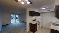 This single-floor residence offers a practical and comfortable layout with an open-concept design that connects the kitchen and living area seamlessly. The kitchen is equipped with dark espresso-toned cabinetry that provides ample storage, complemented by light-colored countertops, a white electric range, and a full-size refrigerator. The overhead cabinetry extends above the range, maximizing vertical storage space. Tile flooring in the kitchen transitions into plush carpeting in the living area, creating a warm and inviting atmosphere. The living space is illuminated by a ceiling fan with a light fixture, adding both function and character to the room. A sliding glass door at the far end of the living area floods the space with natural light and provides direct access to the outdoors, offering views of the exterior surroundings. The textured ceiling adds a subtle architectural detail throughout. Overall, the space presents a clean, functional design with a neutral color palette that offers a versatile canvas for personalization.