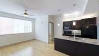 This well-designed single-floor unit offers a contemporary open-concept layout that seamlessly connects the kitchen and living areas. Light hardwood flooring runs throughout, brightening the space and lending a clean, modern aesthetic. The kitchen is a standout feature, showcasing rich dark cabinetry, granite countertops, and a breakfast bar peninsula with an integrated sink — ideal for both meal preparation and casual dining. Black appliances, including a range and built-in microwave, complement the kitchen's bold, contrasting color palette. Pendant lighting hangs above the peninsula, adding both functionality and a decorative touch. The adjoining living area benefits from a ceiling fan and a large window dressed with horizontal blinds, allowing for natural light while maintaining privacy. Crisp white walls throughout the space enhance the sense of openness and keep the overall atmosphere feeling airy and fresh. A hallway visible from the main living area leads to additional rooms, suggesting a thoughtful and practical floor plan. The space strikes a balance between modern style and everyday functionality, making it a comfortable and visually appealing environment.