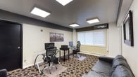 This single-floor office space is designed for productivity and professional comfort. The room features two glass-top desks, each paired with ergonomic mesh chairs, creating a practical dual-workstation setup ideal for collaborative or independent work. Each station is equipped with computer monitors, and a shared printer sits conveniently between them. The space is anchored by a decorative area rug with a bold floral pattern, adding warmth and character to the room. Overhead, a dark-painted ceiling hosts multiple large flush-mount LED panel lights that provide bright, even illumination throughout the workspace. A wall-mounted mini-split air conditioning unit ensures year-round climate control. A large window fitted with horizontal blinds allows for natural light while maintaining privacy. Baseboard heating runs along the exterior wall, complementing the HVAC system. A framed cityscape triptych on the wall adds a stylish, urban aesthetic to the otherwise clean and neutral interior. The overall ambiance is modern and functional, striking a balance between a professional work environment and an inviting, well-designed interior.