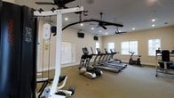 This fitness center offers a comprehensive and well-organized workout environment designed to meet a range of exercise needs. The space is outfitted with multiple treadmills, elliptical machines, and an upright bike, providing ample options for cardiovascular training. A multi-station cable machine anchors one side of the room, supporting a variety of strength and resistance exercises. The flooring throughout is a durable, cushioned surface well-suited for high-traffic gym use. Overhead, two ceiling fans ensure consistent air circulation, complemented by recessed lighting that keeps the space bright and energizing. Wall-mounted flat-screen televisions are positioned throughout the room, allowing members to stay entertained during their workouts. Natural light filters in through several windows, adding an open and welcoming atmosphere to the interior. The neutral-toned walls and clean layout contribute to a focused and distraction-free environment. Overall, this fitness center is thoughtfully arranged to maximize usable floor space while maintaining a comfortable, accessible, and fully functional setting for users of all fitness levels.