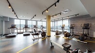 Located at 150 Main St in Hackensack, NJ, this well-appointed fitness center offers a comprehensive workout environment designed for a wide range of exercise needs. The space is defined by its floor-to-ceiling windows that line the perimeter, flooding the interior with natural light and providing open views of the surrounding cityscape.

The facility is organized into distinct workout zones, clearly delineated by bold yellow floor markings. The cardio section is generously stocked with a full lineup of treadmills, upright and recumbent bikes, ellipticals, and rowing machines — all featuring modern digital displays. A dedicated strength training area houses a tiered dumbbell rack with a comprehensive weight selection, multiple flat benches, and a cable/weight machine setup suitable for a variety of resistance exercises.

Structural columns finished in a distinctive yellow accent add a modern industrial character to the space. Exposed brick walls and wood-textured wall panels provide visual contrast alongside the sleek gray painted walls and white ceilings. Track lighting mounted throughout ensures even illumination across all workout areas, complementing the abundant natural light.

The durable rubber flooring throughout provides both safety and comfort during workouts. Large mirrors in the strength training section allow users to monitor form. The overall design is clean, functional, and contemporary, creating an energizing atmosphere well-suited for daily fitness use.