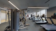 Located at 200 Presidential in Woburn, Massachusetts, this contemporary fitness center offers a well-appointed, single-floor facility designed to accommodate a wide variety of workout routines. The space is organized into distinct training zones, each thoughtfully equipped to serve different fitness needs.

The cardio area lines an expansive wall of floor-to-ceiling windows, allowing abundant natural light to fill the space and providing views of the surrounding landscape. Treadmills, elliptical trainers, and rowing machines are arranged to take full advantage of these outdoor sightlines. Sleek linear LED lighting runs along the ceilings throughout, complementing the modern, gray-toned aesthetic.

The strength training zone features a multi-station cable machine, a squat rack with bench, free weights, and resistance equipment. A decorative patterned accent wall adds visual interest to this area, while patterned carpet flooring provides both functionality and style.

A dedicated boxing and functional training zone stands out with its bold red-and-white heavy bag stations, a mini trampoline, and an organized rack of medicine balls and training accessories. Full-length mirrors amplify the sense of space in this section. The facility also includes additional strength machines, a wall-mounted display, and private rooms accessible from the main floor, rounding out this comprehensive fitness environment.