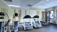 This fitness center is thoughtfully designed to accommodate a range of workout needs within a single-floor layout. The space is divided into distinct zones, each serving a specific training purpose. The cardio area is a central highlight, housing multiple treadmills and an elliptical machine arranged in a row, allowing users ample space to exercise comfortably. Large wall-mounted mirrors line this section, visually expanding the room and enabling users to monitor their form during workouts.

Adjacent to the cardio zone, a multi-functional cable and weight machine provides options for strength training exercises targeting various muscle groups. A weight bench and additional resistance equipment round out the strength training offerings. A separate area toward one end of the space accommodates free weights and functional fitness equipment, including a stability ball and additional accessories.

The fitness center features a drop ceiling with recessed fluorescent panel lighting, ensuring consistent, well-distributed illumination throughout. Warm beige walls with subtle architectural column detailing give the space a clean, professional aesthetic. Durable rubber-mat flooring provides safety and comfort underfoot. A window on one wall allows natural light to supplement the overhead lighting, contributing to an open, energizing atmosphere conducive to an effective workout experience.