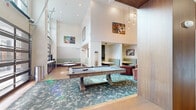 This contemporary communal lounge and dining space is designed to accommodate a variety of social and recreational activities across a single, well-appointed floor. The space is defined by its dramatic double-height ceilings, which amplify the sense of openness and allow natural light to flood in through oversized windows and a striking glass-paneled garage door that opens directly to an outdoor pool area, seamlessly blending indoor and outdoor living.

The main lounge area centers around a full-size pool table set atop a patterned teal area rug, surrounded by plush, channel-tufted velvet armchairs in muted rose and mauve tones. A large dual-screen media wall serves as a focal point, framed by warm wood paneling that extends toward the ceiling. Pendant lighting with Edison-style bulbs adds a warm, ambient glow throughout.

Along one wall, built-in banquette seating upholstered in sage green fabric lines a dining area furnished with round marble-topped bistro tables. A wall-mounted flat-screen television and additional pendant lighting complete this casual dining zone. Private semi-enclosed dining nooks, each equipped with a flat-screen TV, built-in bench seating, and a pendant light, offer intimate spaces for smaller gatherings. Colorful contemporary artwork is displayed throughout, adding a vibrant, curated character to the overall ambiance.