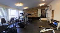 This fitness room offers a well-equipped workout space designed for convenient, on-site exercise. The room is characterized by its exposed brick accent wall, which adds a rugged, industrial aesthetic to the space, complemented by dark rubber flooring that provides durability and traction during workouts. Overhead, flush-mounted square light fixtures ensure consistent, even illumination throughout the room.

Natural light enters through large windows fitted with vertical blinds along one wall, creating a bright and inviting atmosphere. The cardio equipment selection includes elliptical trainers and treadmills positioned toward the center and along the walls, offering users a variety of aerobic workout options. A stationary bike and additional strength or stretching equipment are also present, rounding out the available fitness options.

A large mirror is mounted along one wall, enhancing the sense of space and allowing users to monitor their form during exercise. A railing near the rear of the room suggests a split-level or staircase access point nearby. Overall, the space is functional, cleanly organized, and equipped to support a range of fitness routines.