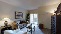 This single-floor residence offers a well-appointed living space designed for comfort and functionality. The living room serves as the heart of the home, featuring neutral warm-toned walls and smooth ceilings that create a bright, airy atmosphere. Natural light flows generously into the space through a large sliding glass door, which opens directly onto an outdoor patio — an ideal extension of the interior living area, furnished for relaxing or dining al fresco.

The living room is thoughtfully laid out to accommodate comfortable seating, centered around a wooden coffee table and area rug that define the gathering space. A framed landscape artwork adorns one wall, adding a decorative focal point, while a tall indoor plant beside the sliding door introduces a touch of greenery and life to the room. The flooring transitions between carpet and hard surface, offering both warmth and practicality.

The patio, visible through the glass door, provides a private outdoor retreat with seating and views of surrounding landscaping. Overall, the space presents a clean, cohesive aesthetic with a welcoming ambiance — practical in its layout while remaining comfortable and livable in character.