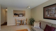 This single-floor residence is thoughtfully laid out to maximize functionality across its living spaces. The open-concept design connects the living, dining, and kitchen areas, creating a cohesive and practical flow throughout the unit.

The living area is comfortably sized, featuring neutral-toned carpeting and warm beige walls that contribute to a welcoming ambiance. A sofa with patterned accent pillows anchors the seating space, complemented by a wooden coffee table and natural greenery that adds a touch of life to the room. Track lighting overhead provides focused illumination.

The kitchen is accessible via a pass-through window that opens directly into the dining area, enhancing the sense of connectivity between spaces. The kitchen features wood-toned cabinetry, standard appliances including a microwave, and ample counter space. Adjacent to the kitchen, the dining area accommodates a compact round wooden table with matching chairs, ideal for everyday meals.

The floor plan also reveals additional rooms branching off the main living space, including what appears to be a bedroom and bathroom, each privately situated. Throughout the residence, neutral finishes, consistent flooring materials, and a practical layout make this a well-organized and livable space.