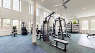 This fitness center is designed to accommodate a wide range of workout routines within a generously proportioned, single-floor layout. The space is architecturally distinguished by soaring double-height ceilings and expansive floor-to-ceiling windows that line the walls, flooding the interior with natural light and offering views of the surrounding landscape. Ceiling fans provide additional air circulation throughout the main workout area.

The cardio zone is well-stocked with multiple treadmills, elliptical trainers, and stair climbers, all positioned to take advantage of the natural light and outdoor views. A dedicated strength training area houses a multi-station cable machine along with various benches and selectorized equipment suited for full-body conditioning. The free weight section features a comprehensive dumbbell rack, kettlebell storage, medicine balls, and stability balls, complemented by rubber flooring for safety and durability.

A secondary area with teal-toned carpet and a decorative accent wall provides additional training space, complete with cable equipment and open floor space for functional movement or stretching. Colorful wall art adds a vibrant, energetic touch to the overall aesthetic. Recessed lighting, white-paneled walls, and mirrored surfaces contribute to a bright, clean, and motivating environment throughout.